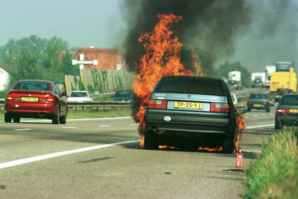 Ein Auto in Flammen auf der Seite einer Straße, mit anderen Fahrzeugen in der Nähe, Bäumen, Gebäuden und einem klaren blauen Himmel im Hintergrund und Gras mit einem Feuerlöscher auf der rechten Seite.