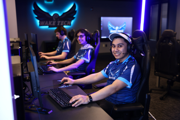 Gruppe von Menschen an einem Schreibtisch mit Computern, Kopfhörern und strahlenden Gesichtern vor Monitoren und Tastaturen, mit einem Logo und Text an der Wand dahinter.