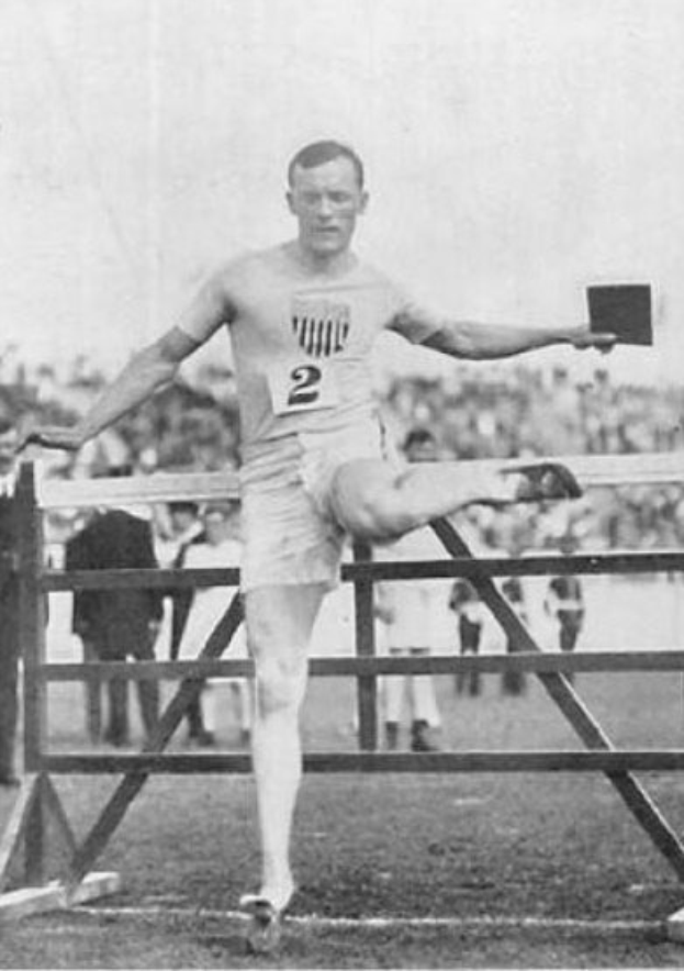 Schwarzes und weißes Foto eines Mannes in einer nummerierten Sportbekleidung, der über ein Hindernis springt, mit Zuschauern im Hintergrund und Himmel darüber.