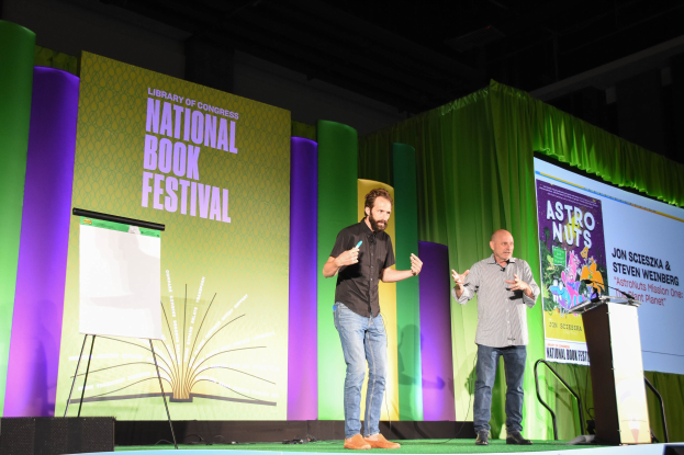 Zwei Männer auf einer Bühne beim Nationalen Buchfestival mit einem Rednerpult und einem Mikrofon rechts daneben und einem großen Bildschirm dahinter.