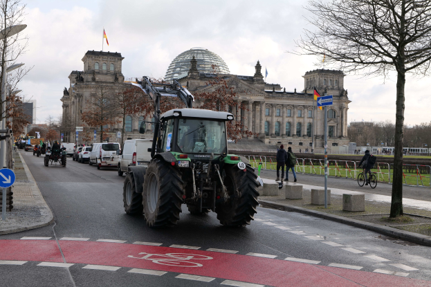 Ein Traktor fährt eine Straße entlang, die von Bäumen, Laternenpfählen und Schildern gesäumt ist, vor dem Reichstaggebäude in Berlin, Deutschland, mit Menschen, die zu Fuß und Fahrrad fahren, auf dem Gehweg und dem Gebäude, das mit Flaggen geschmückt ist, unter einem bewölkten Himmel.