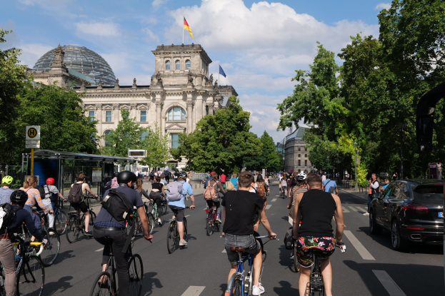 Eine Gruppe von Menschen, die auf Fahrrädern eine von Bäumen gesäumte Straße in Berlin, Deutschland, entlangfahren. Im Hintergrund sind Gebäude zu sehen und auf der rechten Seite ist eine Bushaltestelle zu erkennen.