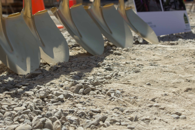 Eine Reihe von Schaufeln auf einem Schotterfeld mit verstreuten Steinen und einem baubezogenen Schild und Verkehrskegel im Hintergrund.