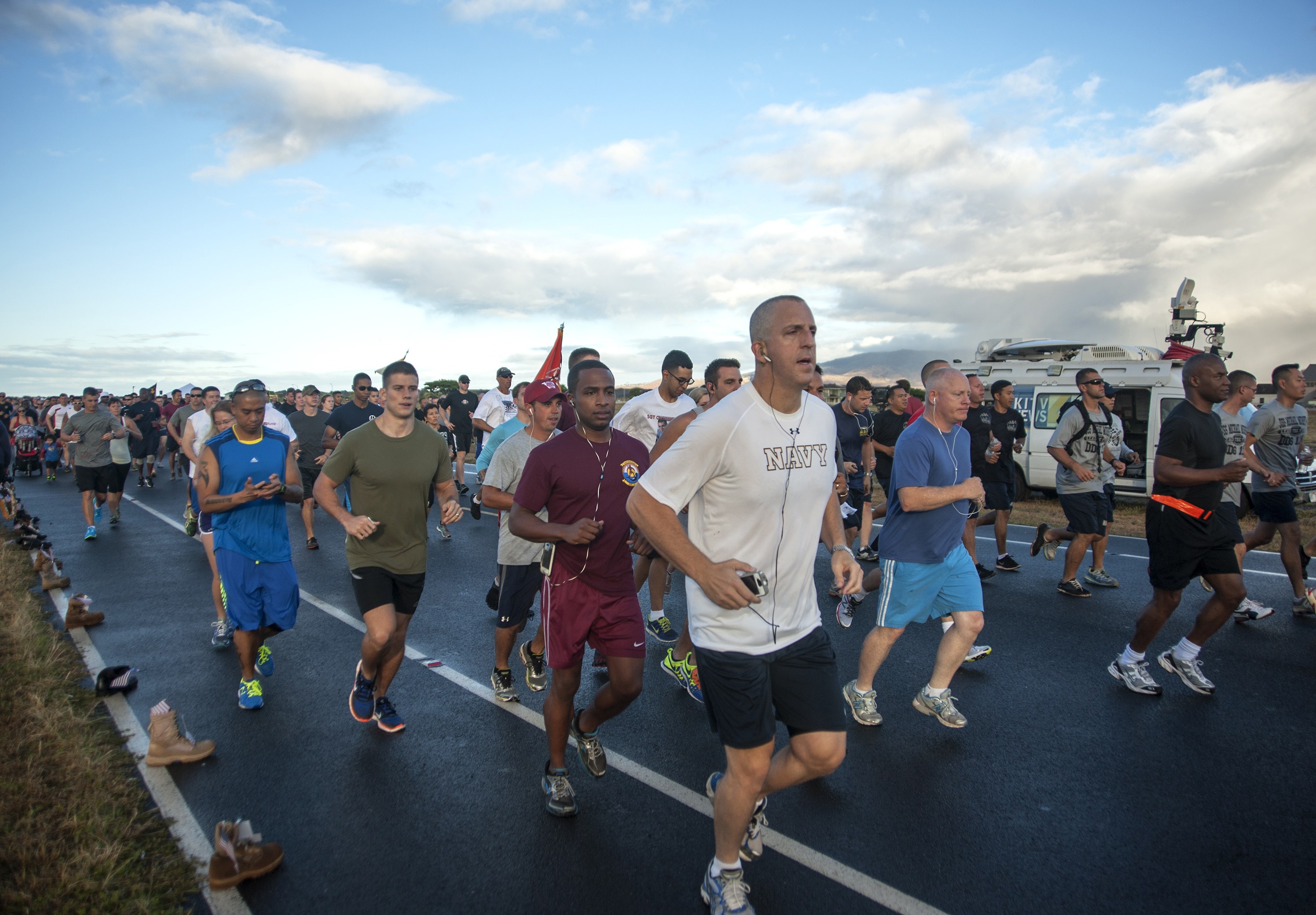 Gruppe von Menschen, die bei einem Marathon auf einer Straße mit Gras auf beiden Seiten laufen, tragen sportliche Schuhe, mit Fahrzeugen, Bäumen, Hügeln und einem bewölkten Himmel im Hintergrund.