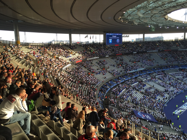 Große Zuschauermenge in einem Stadion bei einem Fußballspiel mit einer Bühne, Fahnen, Stangen und einem Bildschirm im Hintergrund.