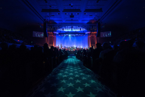 Ein großes Auditorium mit sitzenden Zuschauern, eine Bühne mit Musikern und über Kopfbeleuchtung und Bildschirme, was auf ein Konzerthaus während der Nationalkonvention 2019 hinweist.