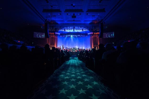 Ein großes Auditorium mit sitzenden Zuschauern, eine Bühne mit Musikern und über Kopfbeleuchtung und Bildschirme, was auf ein Konzerthaus während der Nationalkonvention 2019 hinweist.