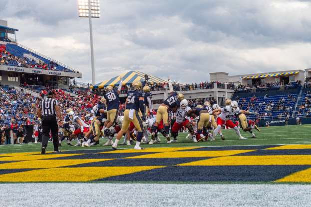 Fußballspieler in Sportkleidung und Helmen konkurrieren auf einem Feld mit Zuschauern, Stadionstrukturen und Text unten.