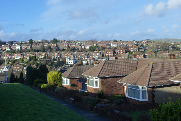 Luftaufnahme eines Wohngebiets mit Häusern, Grünflächen, einer Straße, Strommasten und einer bewölkten Himmel.