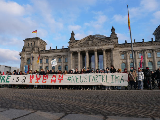Gruppe von Menschen vor dem Reichstaggebäude in Berlin mit einer Fahne, auf der "Wir sind ein Menschenrecht" steht, zu sehen. Im Hintergrund sind Flaggen und Wolken am Himmel zu erkennen.