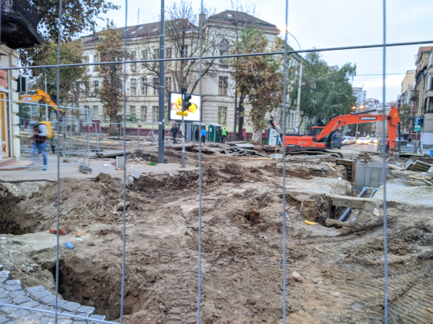Baustelle mit einem großen Loch, umgeben von einem Metallzaun, Fahrzeuge auf der Straße, Fußgänger auf dem Gehweg, Verkehrsampeln, Schilder, Bäume, Gebäude mit Fenstern und einen klaren blauen Himmel.