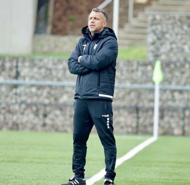 Ein Fußballtrainer mit verschränkten Armen in einem schwarzen Jackett und Hose vor einem Zaun, Treppe und Wand während eines Trainings.