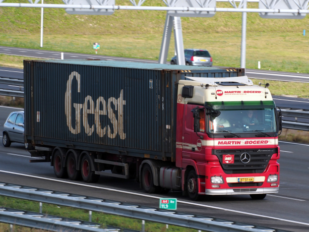 Ein roter Lastwagen mit einem Container auf dem Rücken fährt auf einer Autobahn, mit ein paar Menschen darin und Polen, Brettern und Gras im Hintergrund.