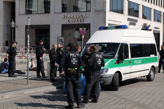 Polizeibeamte vor einem Starbucks-Café mit einem Van auf der rechten Seite und Menschen auf der linken Seite, Hintergrund enthält ein Gebäude mit Fenstern, ein Schild, einen Laternenmast und eine Absperrung.