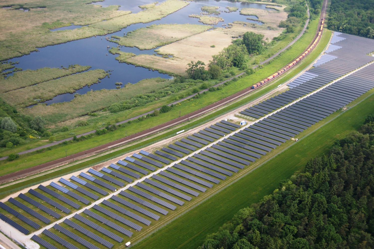 Luftaufnahme einer Solar-Farm mit Solar-Panelen, umgeben von Bäumen, Gras, Pflanzen und Wasser, mit einem Zug auf einer nahen Bahnschiene.