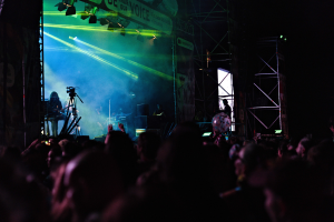 Eine Menge schaut einer Band zu, die auf einer hell erleuchteten Bühne mit Musikinstrumenten, Mikrofonen und einem Banner im Hintergrund spielt.