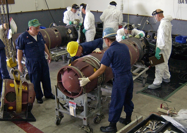Mannergruppe in Helmen und Handschuhen beim Entschärfen einer großen Bombe in einer industriellen Fabrikumgebung mit Maschinen und einer Wandtexttafel im Hintergrund.