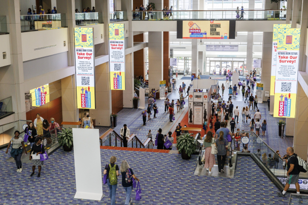 Eine große Gruppe von Menschen geht durch ein großes Gebäude mit Bannern, Topfpflanzen, Rolltreppen, Geländern, Säulen, Texttafeln und Glastüren, einige tragen Taschen, wahrscheinlich nehmen sie am Nationalen Buchfestival teil.