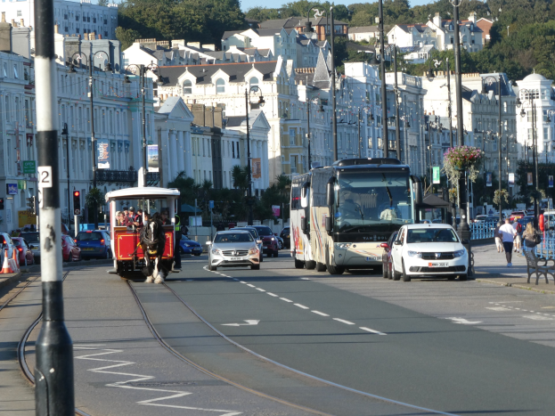 Eine belebte Stadtstraße mit hohen Gebäuden, mehreren Fahrzeugen, Fußgängern auf dem Gehweg, Laternen, Verkehrsampeln, Straßenschildern, Bäumen und einem klaren blauen Himmel.