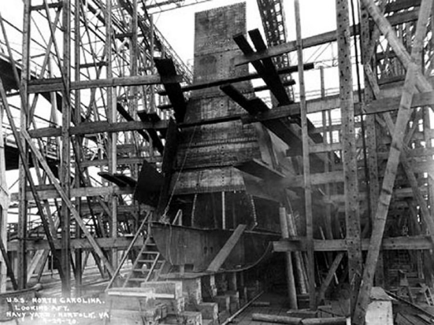 Schwarzes und weißes Foto eines Schiffs in einem Schiffbau, mit Holzpfählen und Treppen im Vordergrund und Himmel im Hintergrund, mit dem Text "Marinebewaffnung" in der linken unteren Ecke.
