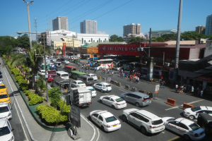 Eine belebte Stadtstraße mit Autos, Bussen, Fußgängern, Bäumen, Straßenlaternen, Strommasten, Gebäuden und einem klaren blauen Himmel.