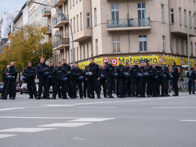 Eine Gruppe von Polizisten in schwarzen Uniformen mit blauen Helmen steht in einer Straße, die von Laternenmasten, Bäumen und Glasfenstern gesäumt ist, unter einem klaren blauen Himmel.