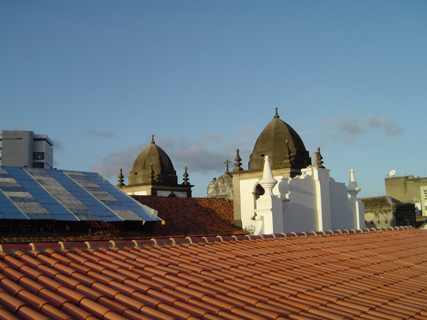 Stadtpanorama mit Gebäuden im Vordergrund und einem blauen Himmel im Hintergrund, mit Solarpanelen auf einem Dach, die den Einsatz erneuerbarer Energie anzeigen.
