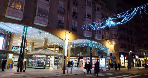 Eine belebte Stadtstraße bei Nacht, beleuchtet von Straßenlaternen und Weihnachtslichtern, mit Menschen, die gehen, Gebäuden mit Fenstern und Balkonen, Geschäften mit Gegenständen, Schildern und Straßenlaternen mit Lichtern unter einem dunklen Himmel.