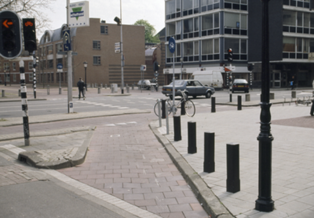Eine Straßenszene in einer Stadt mit einem geparkten Fahrrad, fahrenden Fahrzeugen, Fußgängern, Pfählen, Verkehrszeichen, Schildern, Bäumen, Gebäuden mit Fenstern und einem klaren blauen Himmel.