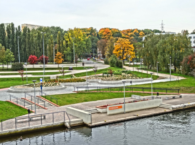 Ein Fluss fließt durch einen grünen Park, der von hohen Gebäuden umgeben ist, mit Bäumen, Straßeninfrastruktur, Fahrzeugen und einem bewölkten Himmel.