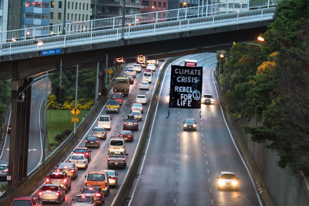 Eine städtische Straße mit starkem Verkehr, eine Brücke im Hintergrund, Bäume, Pfosten, Straßenlaternen, Schilder und Gebäude auf der rechten Seite und ein Banner in der Mitte der Straße.