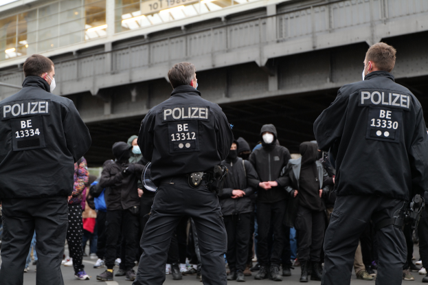 Eine Gruppe von Polizisten in Uniform steht vor einer Menge von Menschen in schwarzen Uniformen und Masken, mit einer Brücke und einem Gebäude im Hintergrund, was auf eine städtische Umgebung während einer Demonstration hinweist.