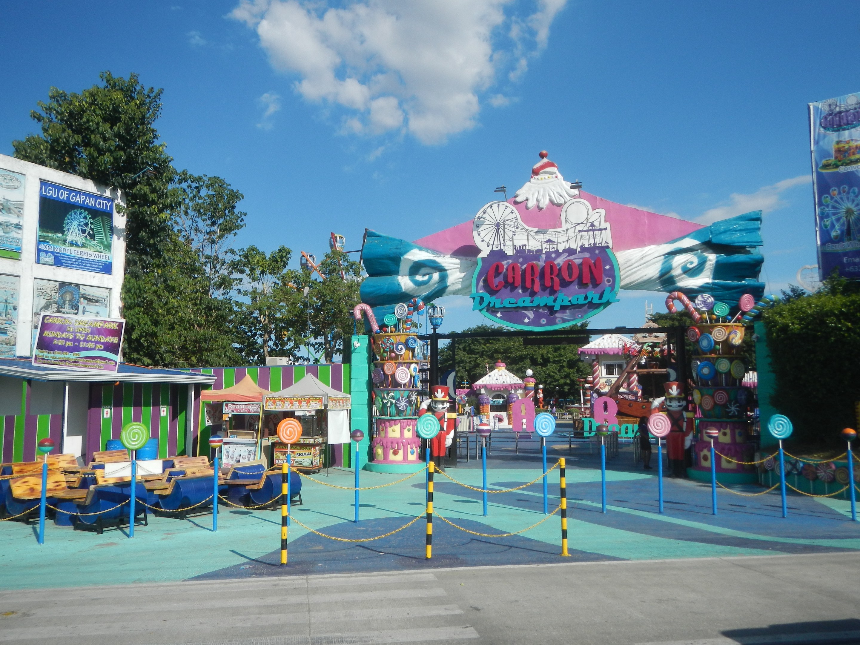 Kirmesfahrgeschäft am Parkeingang mit Straße, Absperrung, Buden, Banner, Bäumen und bewölktem Himmel im Hintergrund.