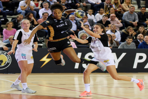 Eine Gruppe von Frauen, die Handball auf einem Platz spielen, mit einem Ball in der Mitte der Aktion, während Zuschauer im Hintergrund sitzen und ein Banner mit Text und Logos sichtbar ist.