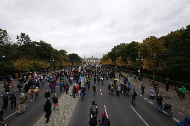 Eine große Gruppe von Menschen marschiert auf einer baumbestandenen Straße in Berlin, hält Kameras, mit einem Gebäude und einem klaren Himmel im Hintergrund.