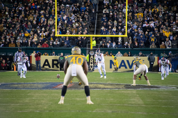 Ein Fußballspiel im Gange mit Spielern in Helmen und Sportbekleidung auf dem Feld und einer Zuschauermenge im Hintergrund, mit sichtbaren Pfählen und Texttafeln.