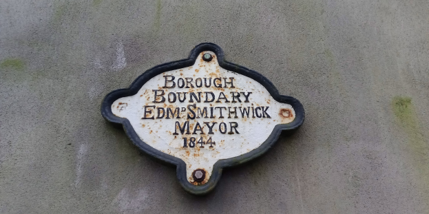 Metallplakette an einer Gebäudeseite mit schwarzem Hintergrund und weißer fettgedruckter Schrift, die "Gemeindegrenze Edm Smithwick Bürgermeister" liest.