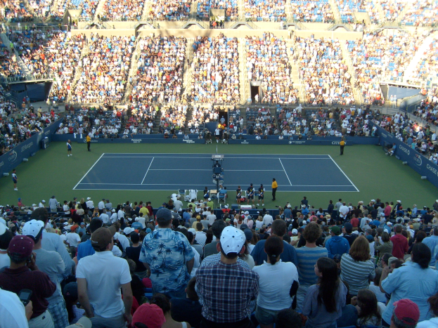 Eine große Menschenmenge schaut einem Tennismatch in einem vollen Stadion zu, mit Spielern auf dem Platz und Zuschauern in den Rängen.