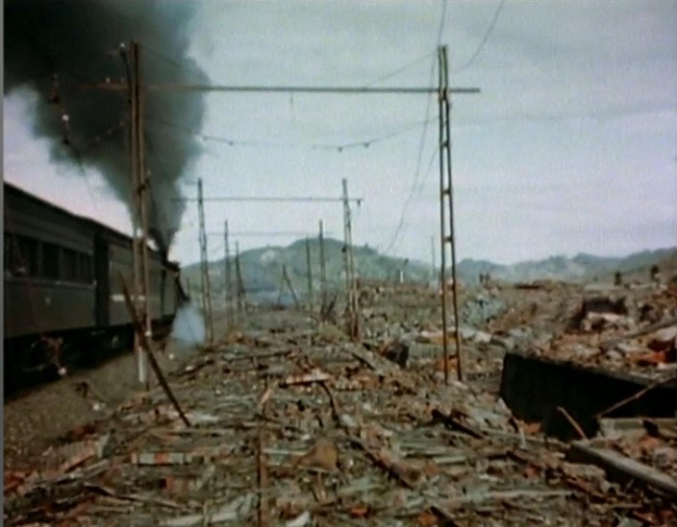 Ein Zug fährt durch ein beschädigtes Gebiet mit Rauch, der aus ihm aufsteigt, umgeben von Strommästen und -drähten, vor Höhen unter einem klaren Himmel, mit verstreuten Trömmern im Vordergrund.