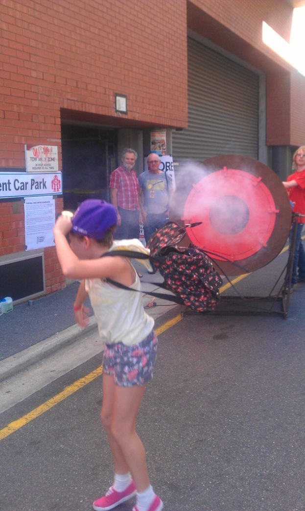 Ein Kind mit Rucksack und Mütze steht auf einer Straße mit Rauch im Hintergrund, ein kreisförmiges Objekt auf einem Ständer, Menschen, Wände, Pfosten auf der linken Seite und Gegenstände auf dem Gehweg.