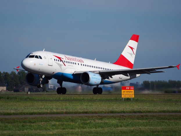 Austrian Airlines Airbus A320-200 startet vom Frankfurt Airport mit einer Tafel im Vordergrund, Gras, Bäumen, Gebäuden und einem klaren blauen Himmel im Hintergrund.