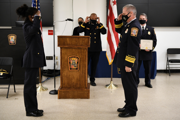 Gruppe uniformierter Personen in Masken vor einem Podium mit Mikrofon, umgeben von Fahnen und Möbeln, während einer Vereidigung eines neuen Feuerwehrchefs