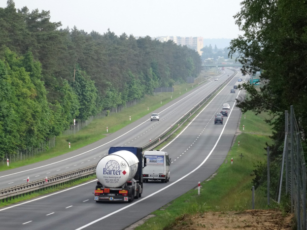 Ein großer Lkw fährt auf einer von beiden Seiten mit Bäumen gesäumten Autobahn, mit einem Zaun und Gras auf der rechten Seite, Gebäuden in der Ferne und einem klaren blauen Himmel darüber.