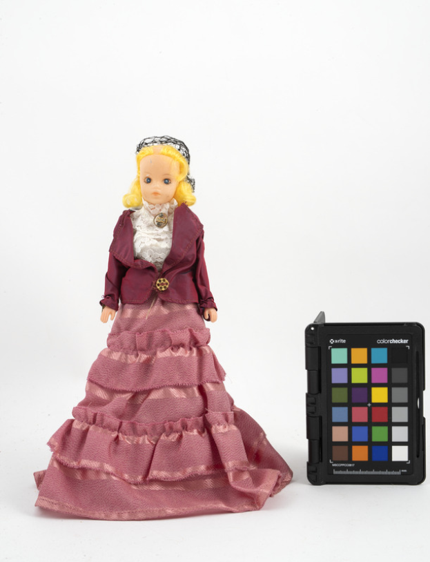 Eine Barbie-Puppe in einem pinken Kleid und einer maronenfarbenen Jacke steht neben einem schwarzen Farbpaletten.