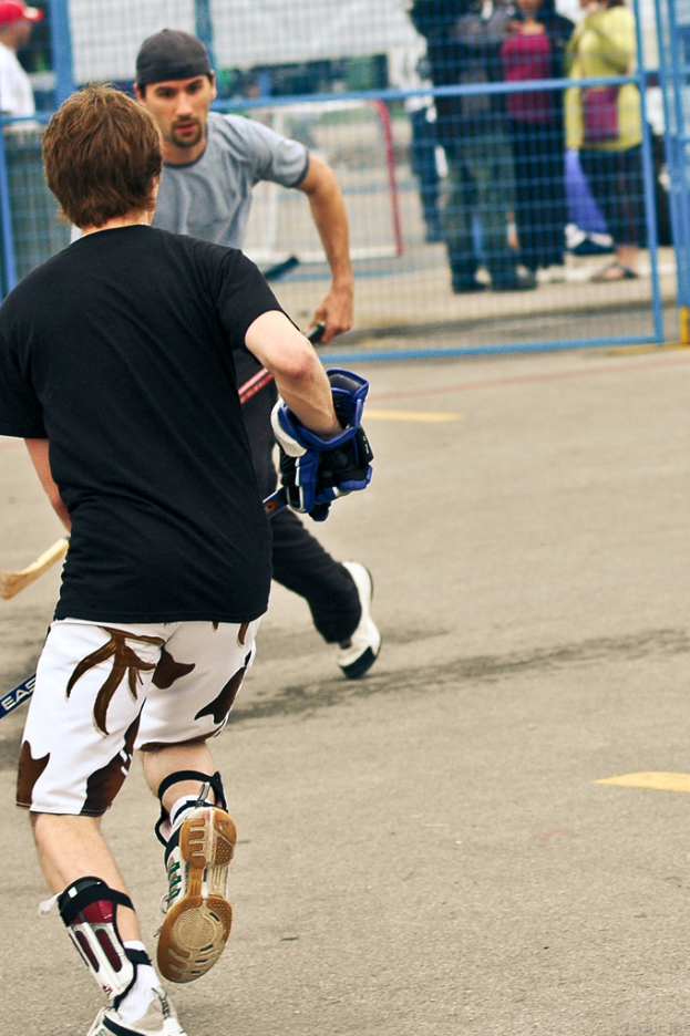 Zwei Personen, die Hockey auf einer Straße mit einem Zaun dahinter und einer Menge Menschen im Hintergrund spielen.