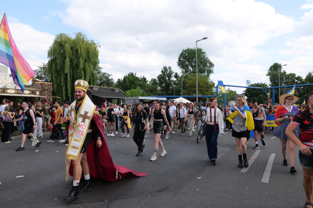 Eine Gruppe von Menschen, die bei der Gay Pride Parade 2018 marschieren und eine Regenbogenflagge sowie Musikinstrumente tragen, mit Laternenmasten, Bäumen, Hütten und einem bewölkten Himmel im Hintergrund.