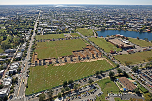 Luftaufnahme einer Stadt mit Gebäuden, Bäumen, Gras, Straßen mit Fahrzeugen, Wasser und Wolken, die einen großen zentralen Platz als Parkfläche zeigt.