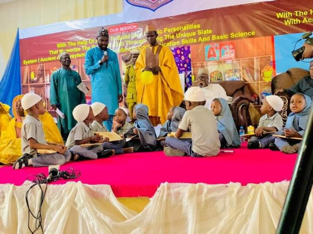 Kinder auf einer Bühne sitzend, die Bücher halten, mit einem Redner am Mikrofon und einem Banner dahinter, während eines Events an einer islamischen Schule in Nigeria.