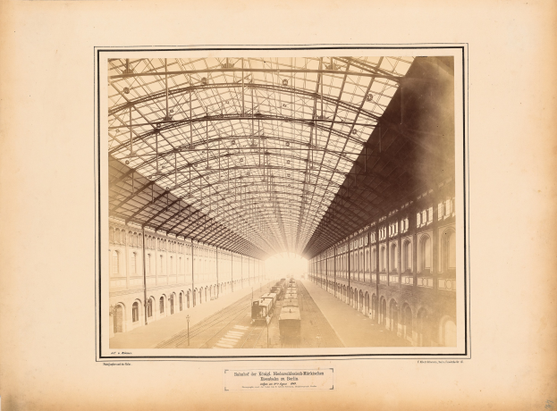 Altes Schwarz-Weiß-Foto eines Bahnhofs mit einem Zug auf den Schienen, flankiert von mehrstöckigen Gebäuden mit Fensterreihen und einem Dach, das von Metallpfosten gestützt wird; verblasster Text erscheint entlang der unteren Kante.