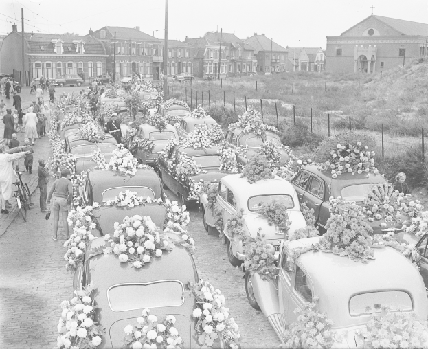 Ein Schwarz-Weiß-Bild einer Trauerprozession mit Autos, die mit Blumenbouquets geschmückt sind, Menschen mit Fahrrädern und Gebäuden im Hintergrund.